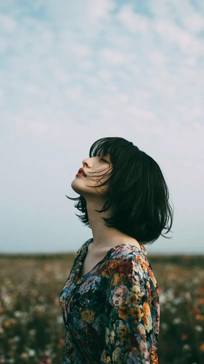 电影极简风格人像：一位美丽的年轻东亚女性独自站立在一片开阔的野花草地上，头顶是无尽的蓝天，她微微仰头，双眼或闭或半睁，仿佛在拥抱风。黑色长发自然蓬松、随风自由拂过面庞，肌肤柔润如…