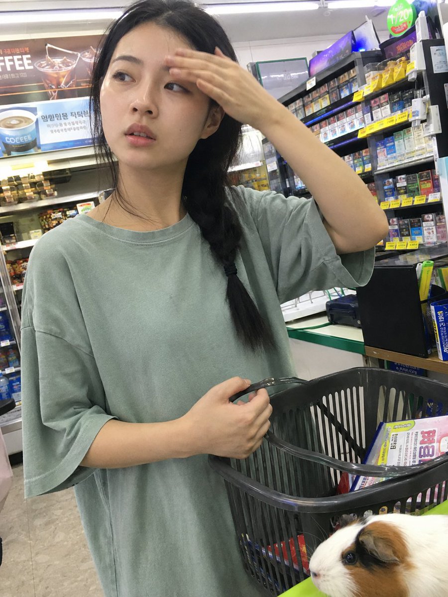 用iPhone前置摄像头意外抓拍的快照，拍摄于一家小型社区便利店内部。一位年轻的韩国女性站在收银台前排队，一边随意地用手抹去额头上的一丝汗珠，一边挎着购物篮…