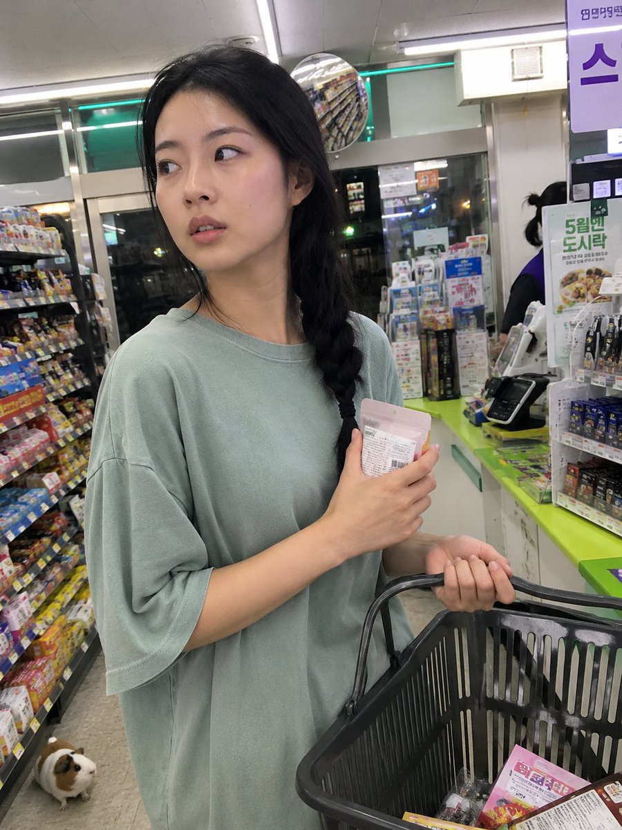 用iPhone前置摄像头意外抓拍的快照，拍摄于一家小型社区便利店内部。一位年轻的韩国女性站在收银台前排队，一边随意地用手抹去额头上的一丝汗珠，一边挎着购物篮。她目光望向一侧，表情…