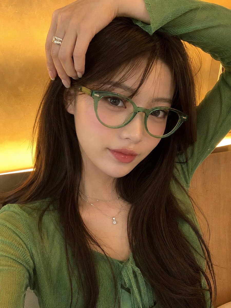 一张高品质的竖版人像照，主体是一位年轻女性，留着长直深棕色头发，戴着时尚的半透明翡翠绿猫眼眼镜，身穿鲜艳的绿色服装。她在室内摆姿势，一只手自然举过头顶，轻轻握着头发。柔和温暖的环…