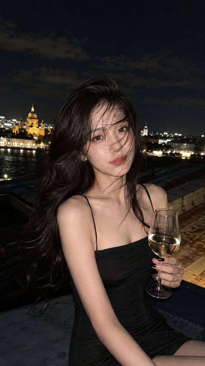 使用参考图像。圣彼得堡屋顶上的半身夜景肖像,一位女性手持一杯白葡萄酒。她身穿修身的黑色吊带连衣裙,露出肩膀,指甲剪短涂成黑色。蓬松有光泽的长发随风飘过面庞。神情平静安详,直视镜头…