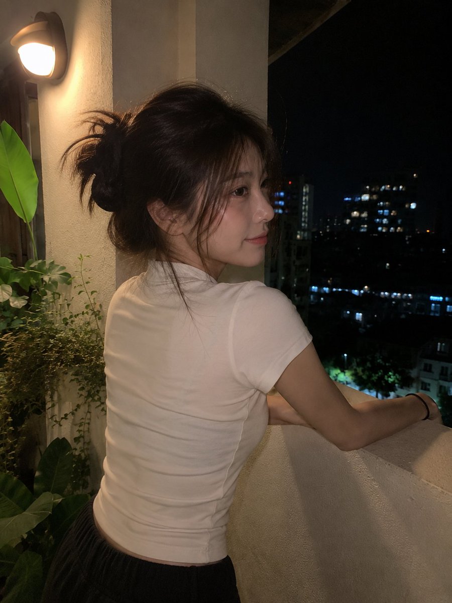 使用 GPT image 2 创建 提示词 一张看起来真实的夜间手机照片：一位年轻的东亚女性随意地站在小型公寓阳台或露台上,由朋友用现代智能手机随手抓拍。她留着中长的深棕色头发…