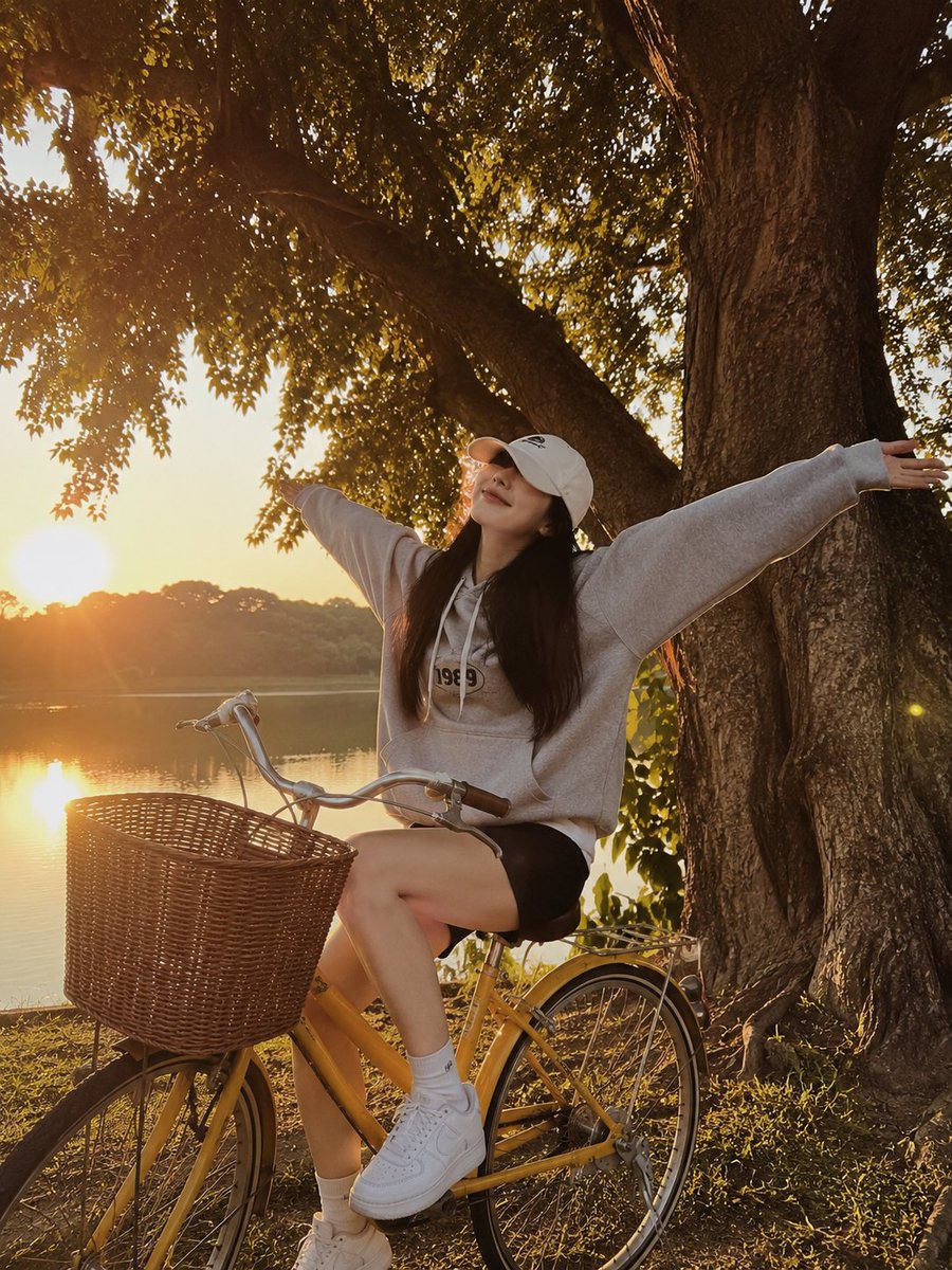一张黄金时段的写实生活方式摄影照片，画面中一位年轻女子俏皮地坐在一辆复古黄色自行车旁，旁边是一片宁静的湖泊。她张开双臂，摆出愉悦、无忧无虑的姿态，嘴角带着柔和的微笑，脸部被一顶白…