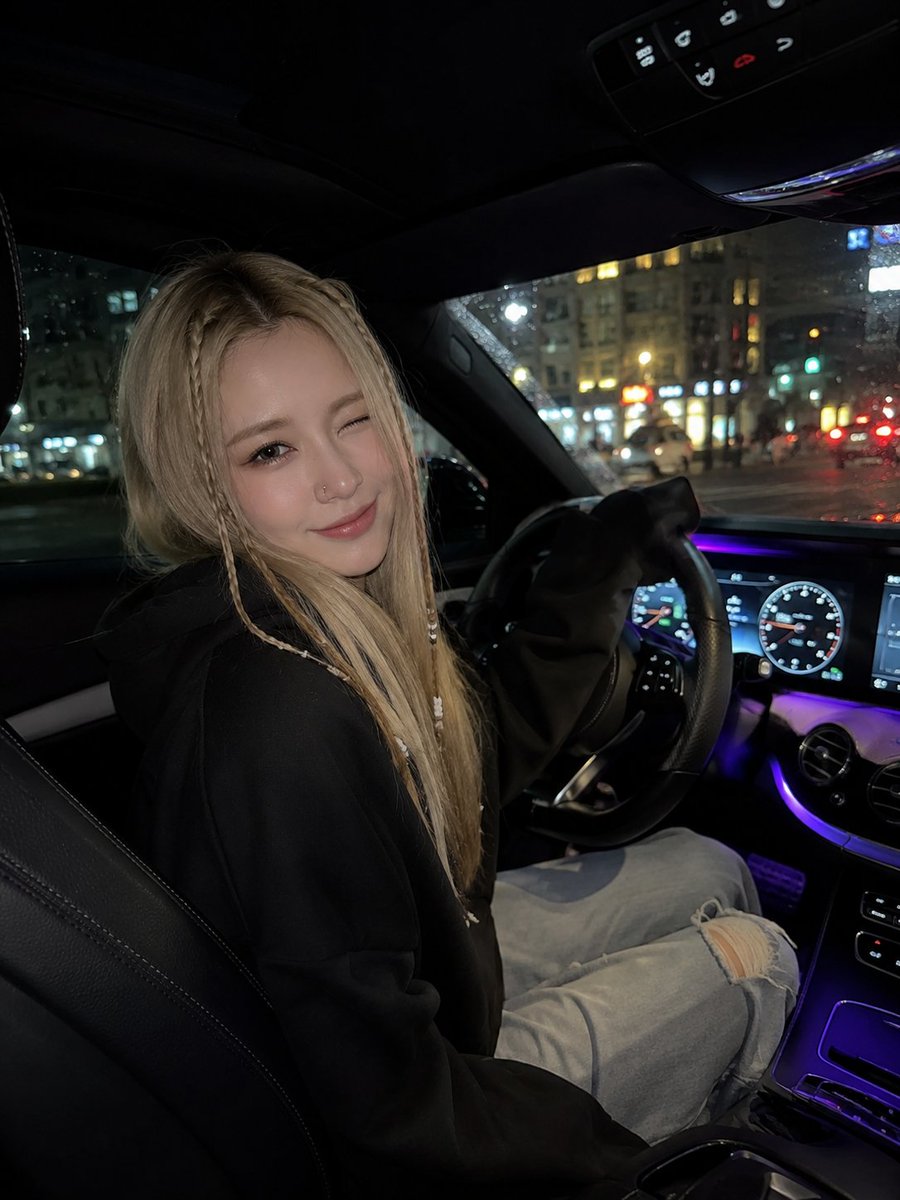 一张超写实的夜间车内自拍，年轻女子坐在驾驶座上，微微转向镜头，调皮地眨眼并带着温柔的微笑。她有一头长直的金发，脸颊两侧各编着精致的细辫，辫子上装饰着小小的白色…