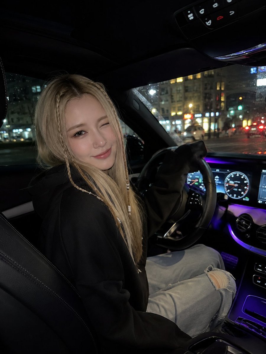 一张照片写实风格的夜间车内自拍照：一位年轻女子坐在驾驶座上，身体微微转向镜头，俏皮地眨着一只眼，露出柔和的微笑。她有一头金色长直发，脸颊两侧各编着几缕精致的细…