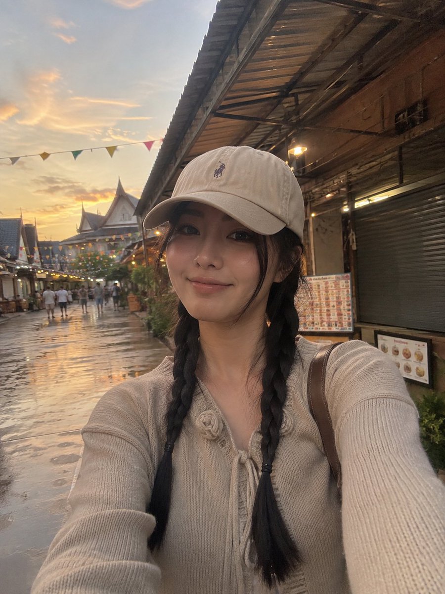 一张照片级写实的竖版旅行自拍照：一位年轻女性在黄金时段身处热闹的集市或旅游村落户外。她戴着米色棒球帽，帽子带有低调的刺绣标志；乌黑长发编成两条整齐的辫子，垂落…