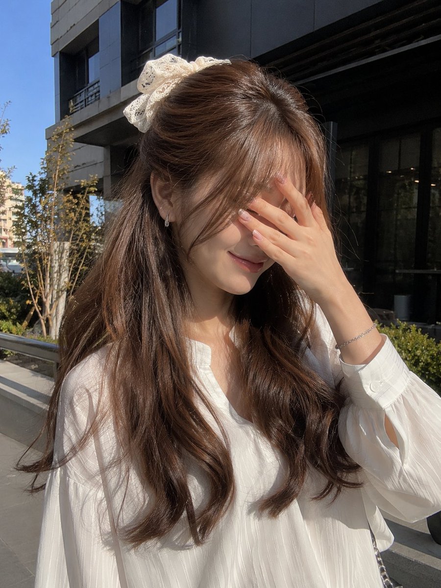 一张年轻女性的户外写实主义肖像照，她站在现代建筑外的明亮自然阳光下。她有一头浓密蓬松的浅棕色长发，呈现出柔和流畅的波浪卷，发帘式刘海轻盈地修饰着面部轮廓。她的…