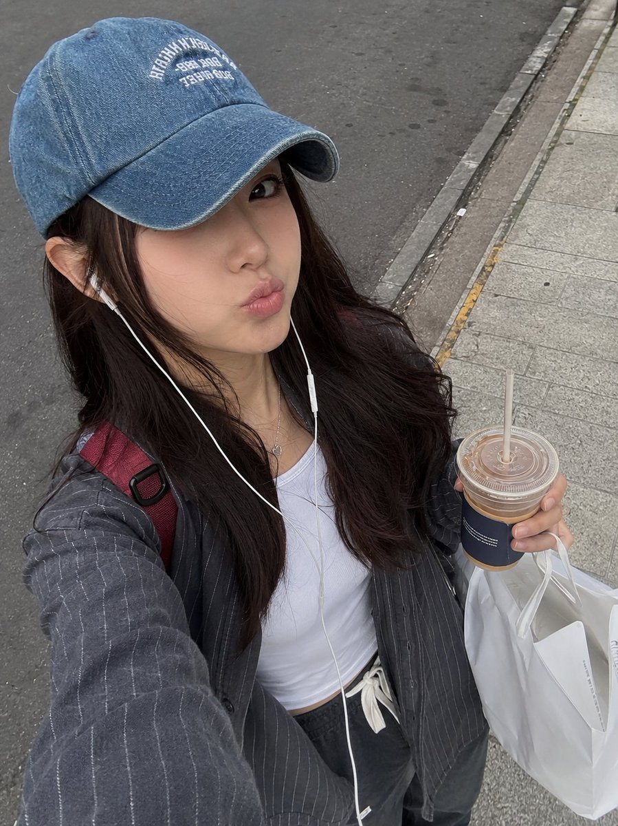 一张照片般逼真的街头风格抓拍自拍照，画面中一位年轻女性正在城市人行道上户外行走。她有一头深棕色长发，自然地垂落在褪色蓝色牛仔棒球帽下，帽子带有白色刺绣字母。她…