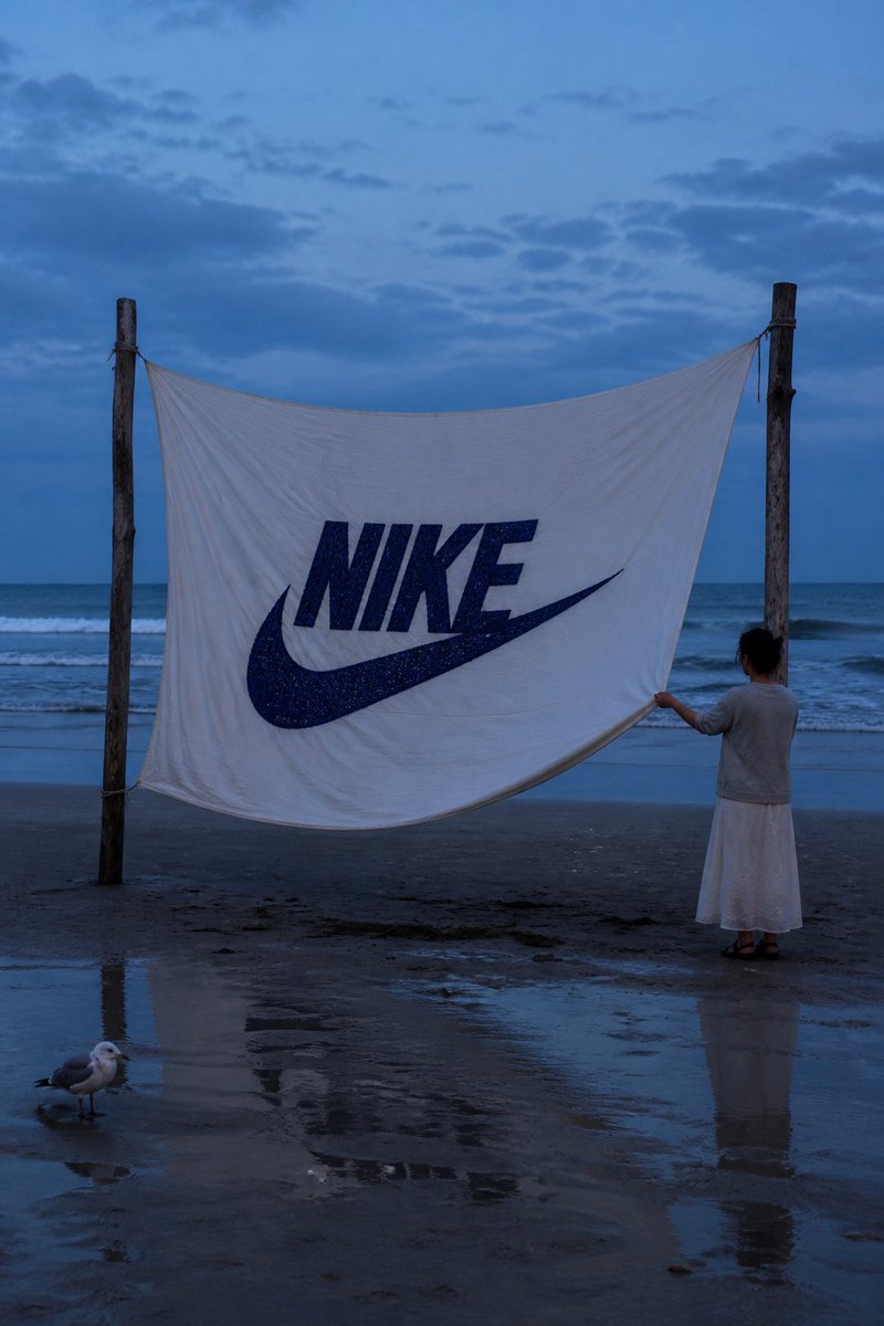 一张写实风格的海岸摄影作品。在蓝调时刻的海滩上，两根浮木杆之间悬挂着一块巨大的白布，中央用粗实的深蓝色线绣有 "NIKE" 字样及其标志，布料随风翻飞飘扬。一…