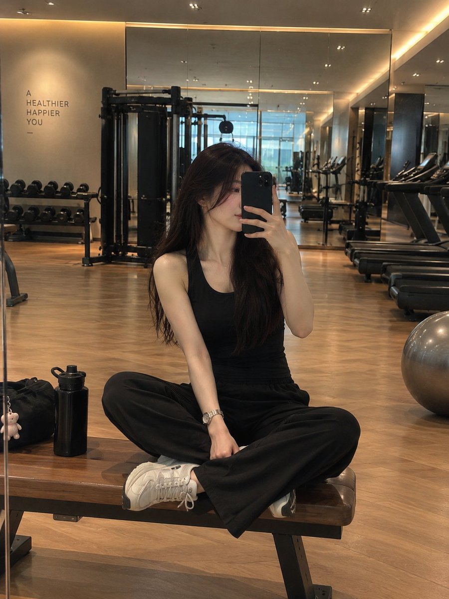 一位时尚年轻女性在现代豪华健身房内对着镜子自拍，盘腿坐在一张木质健身凳上。她留着长而顺直的深棕色头发，神情平静自然，手持一部黑色智能手机置于面前。她身穿一套精…