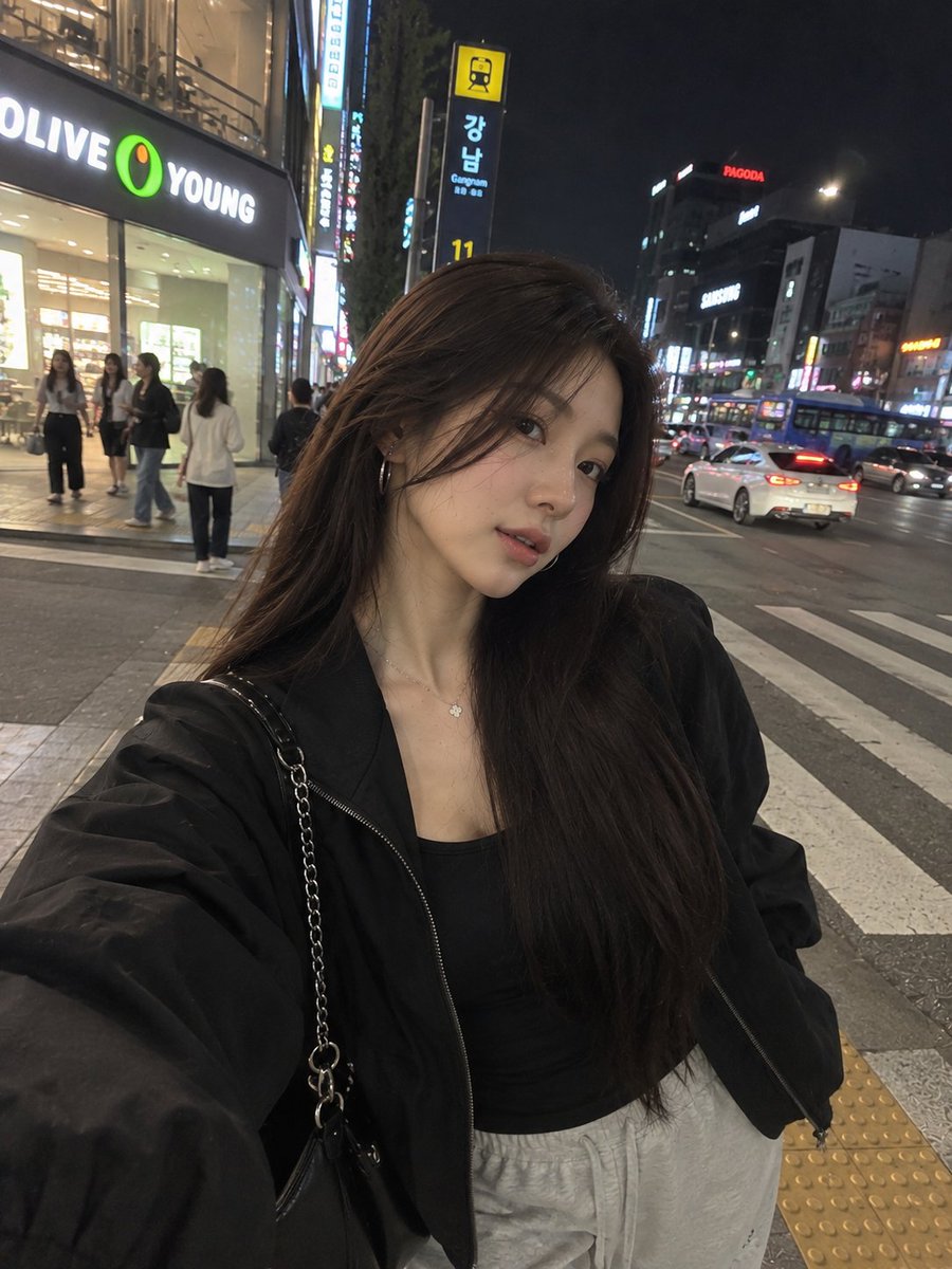 一张照片级写实风格的夜间街头人像照,一位年轻女子在繁华的韩国城市街区中随意地自拍。她留着长而顺直的黑发,妆容柔和自然,佩戴着低调的环形耳环和一条精致的项链。她身穿黑色修身内搭,外…