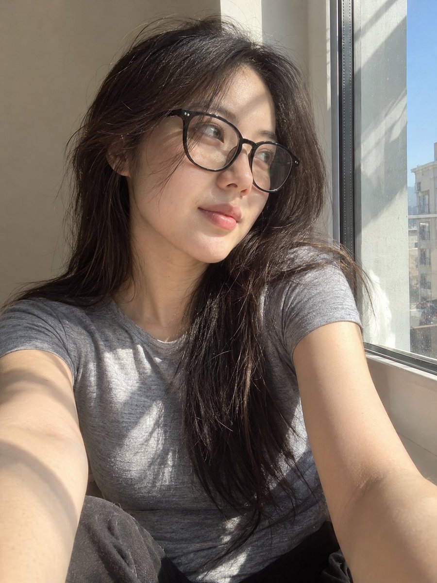 超写实风格的休闲智能手机自拍照：一位年轻东亚女性坐在安静房间内靠近阳光照射的窗户旁。保持与面部参考的高度相似性，包括柔和轮廓的鹅蛋脸、温和的下颌线、自然比例的…