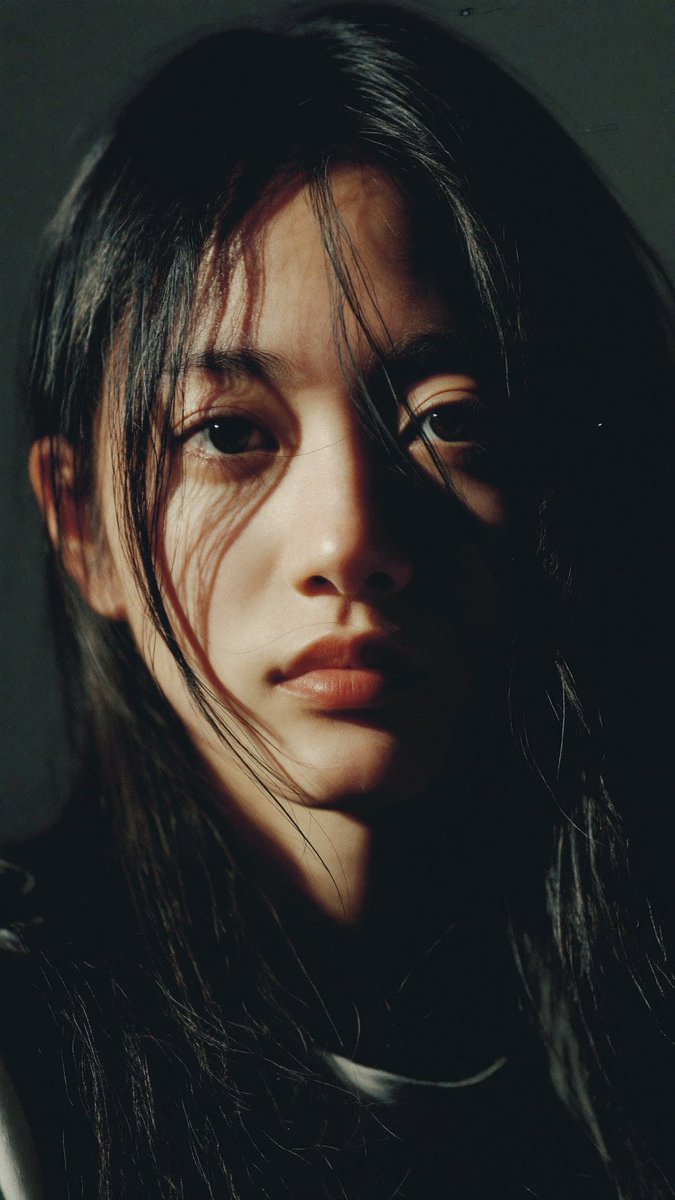 特写电影感人像：一位年轻的东亚女性，长长的略带凌乱的黑发披在肩上，几缕散发拂过脸庞。她直视镜头，表情平静、专注而略带忧郁。左侧柔和而戏剧性的侧光照亮她半边脸庞…