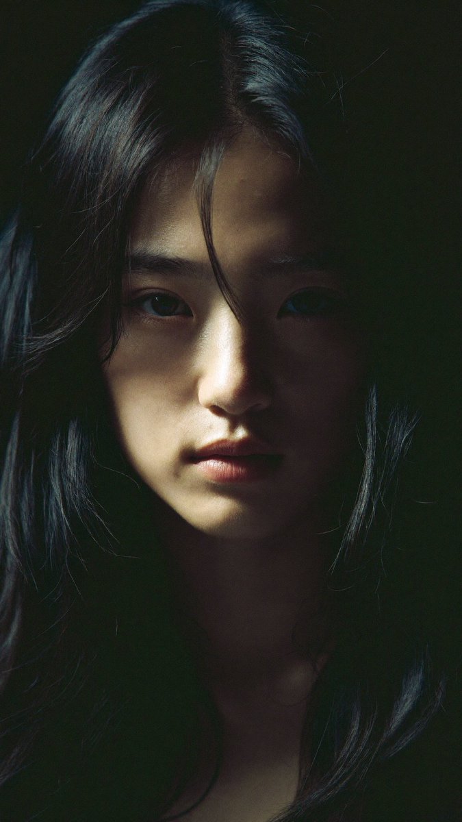 特写电影感人像：一位年轻的东亚女性，长长的略带凌乱的黑发披在肩上，几缕散发拂过脸庞。她直视镜头，表情平静、专注而略带忧郁。左侧柔和而戏剧性的侧光照亮她半边脸庞，另一半则陷入深重的…