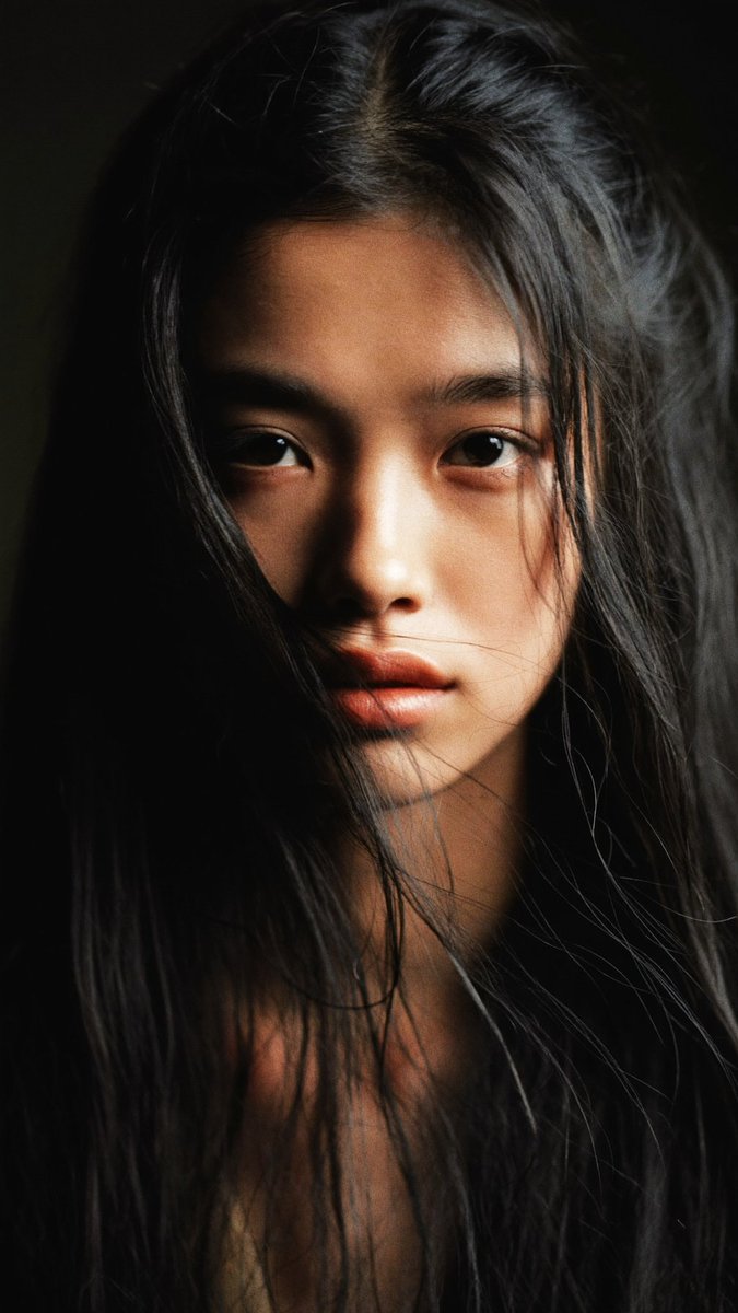 特写电影感人像：一位年轻的东亚女性，长长的略带凌乱的黑发披在肩上，几缕散发拂过脸庞。她直视镜头，表情平静、专注而略带忧郁。左侧柔和而戏剧性的侧光照亮她半边脸庞，另一半则陷入深重的…