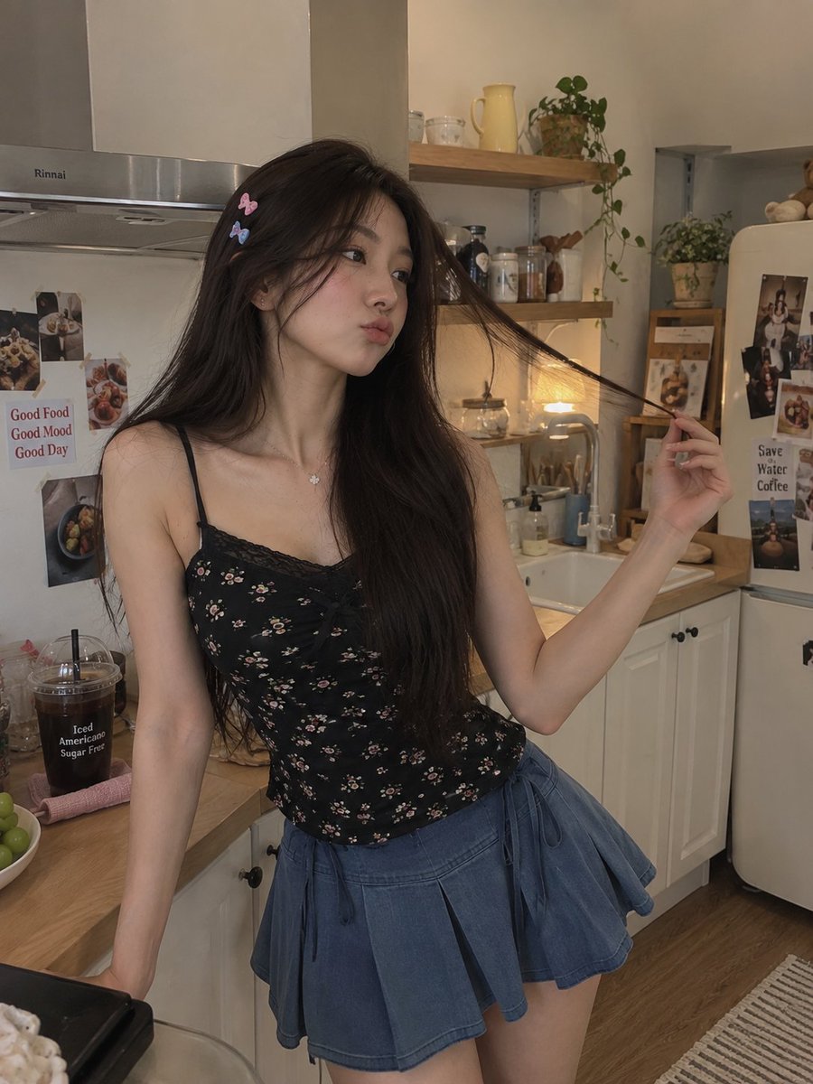 一张高质量的抓拍风格生活人像照,一位年轻女性站在温馨的现代厨房中,身穿黑色碎花细肩带吊带背心和蓝色百褶裙。她有着长而笔直的深棕色头发,别着几枚小巧的彩色发夹…