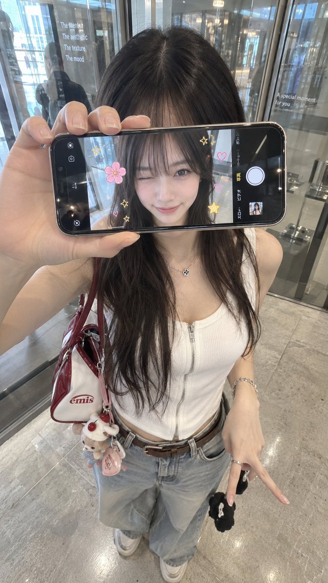 超写实 iPhone 创意人像，身份特征与参考图像完全一致。年轻女性站在室内玻璃店面或反光墙前，从略高的正面角度拍摄。她将一部智能手机水平举在正前方，现实中遮…