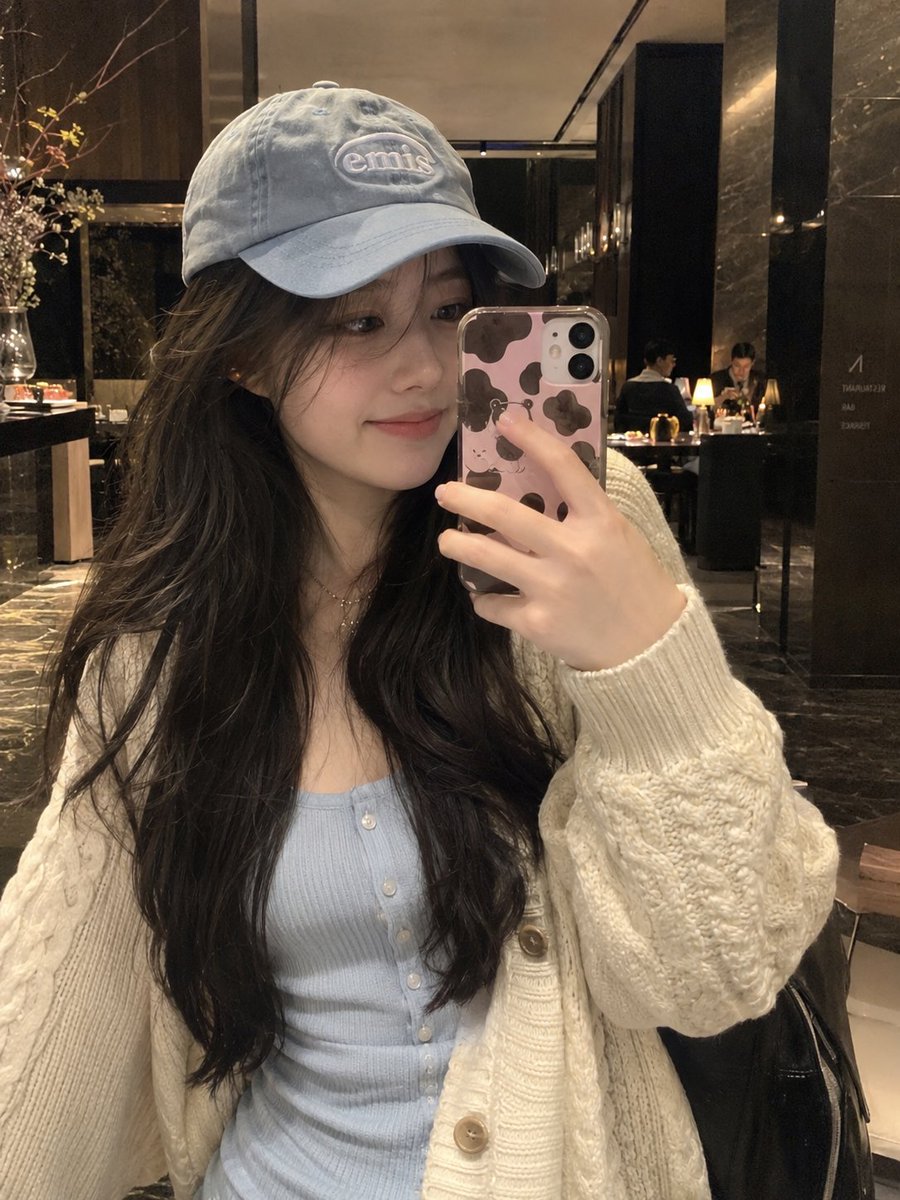 超写实自然智能手机自拍，竖向 3:4，一位年轻东亚女性的室内镜面式随性自拍，背景为一家温馨高档的餐厅或酒店大堂酒廊。 她有着长而自然波浪卷的深棕色头发，披散在…
