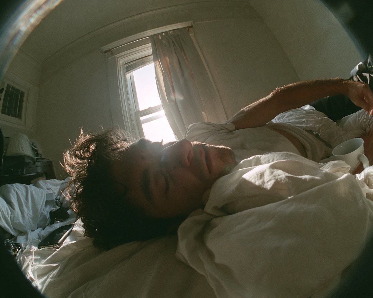 一张鱼眼运动相机风格的抓拍照：一位年轻成年男子刚醒来在床上伸懒腰，强烈的桶形畸变让房间线条弯曲，蓬松的深色头发，朴素的白色床单，晨光透过敞开的窗帘倾泻而入，床…