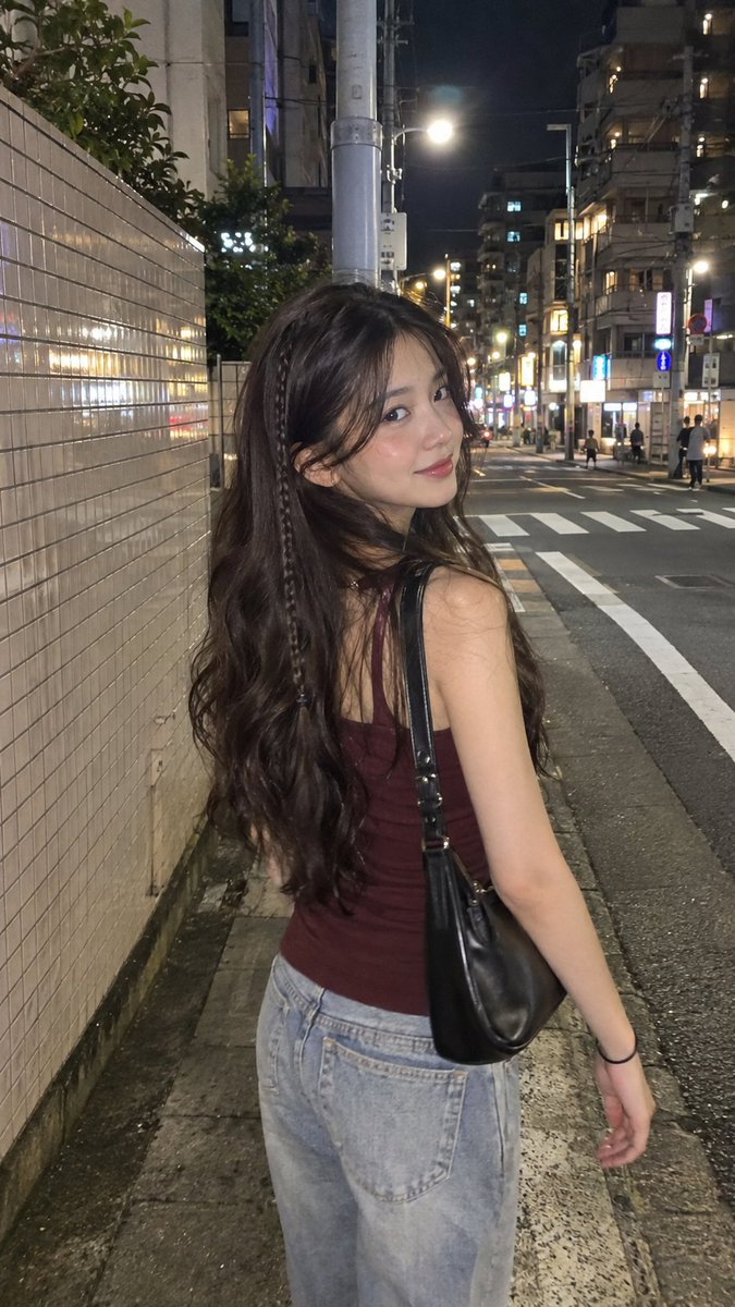 日本夜晚街头的电影感 iPhone 抓拍人像照：一位年轻女性，留着带有细细侧边编发的长波浪深棕色头发，淡妆，嘴角带着若有似无的微笑。她身穿一件修身的酒红色无袖…
